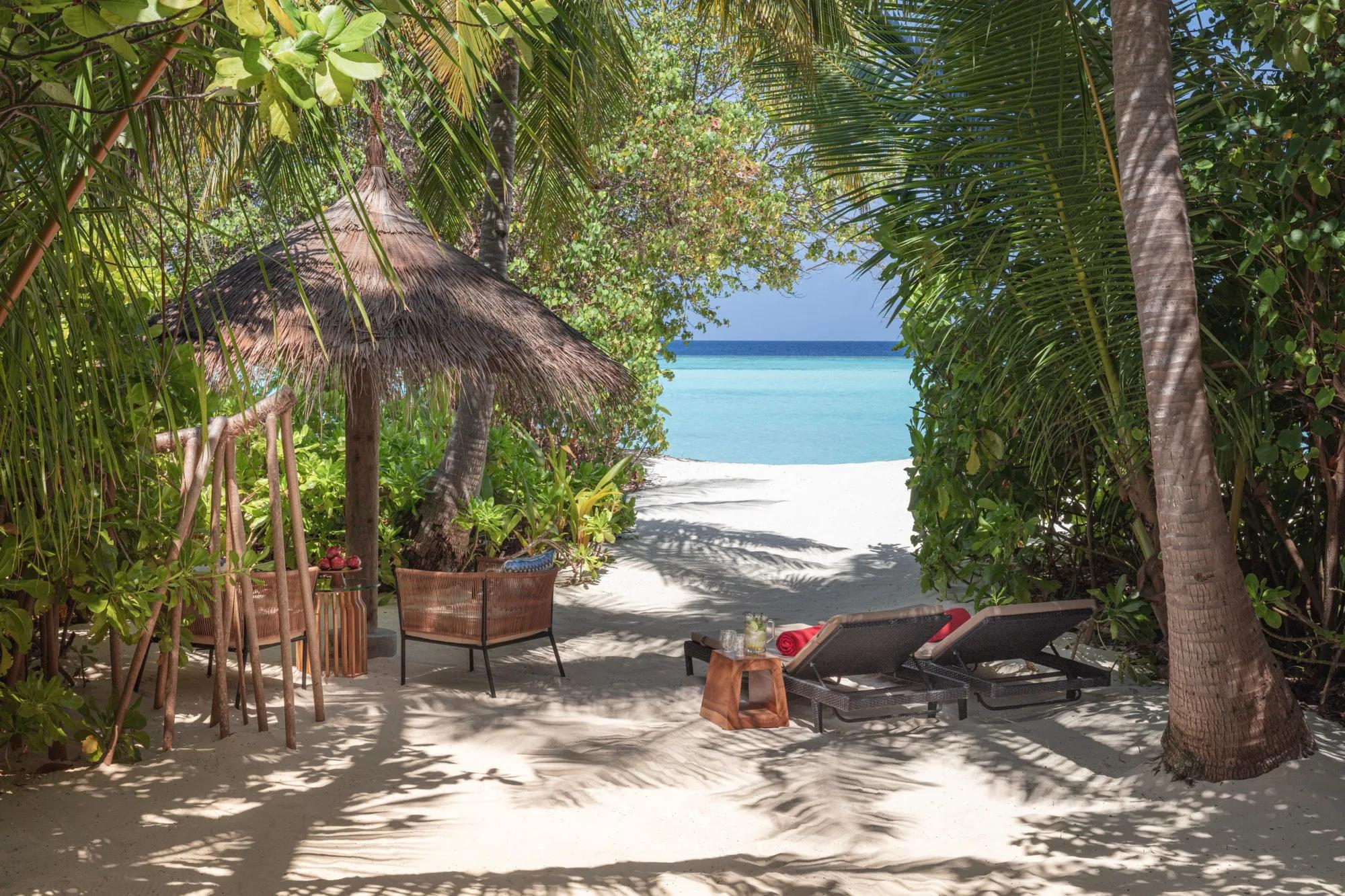 A thatched beach cabana with rattan chairs and loungers, opening through palm fronds onto the lagoon in front of a Sunrise Beach Villa