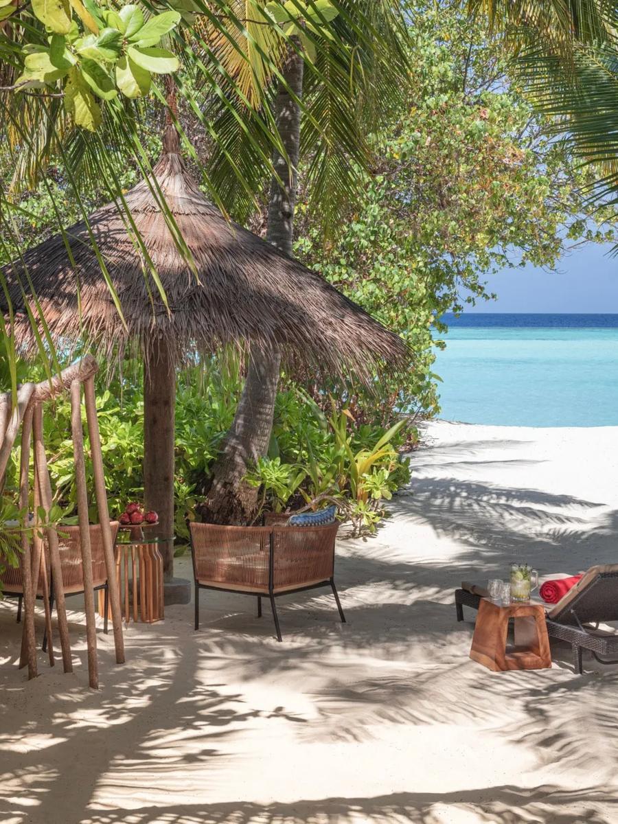A thatched beach cabana with rattan chairs and loungers, opening through palm fronds onto the lagoon in front of a Sunrise Beach Villa