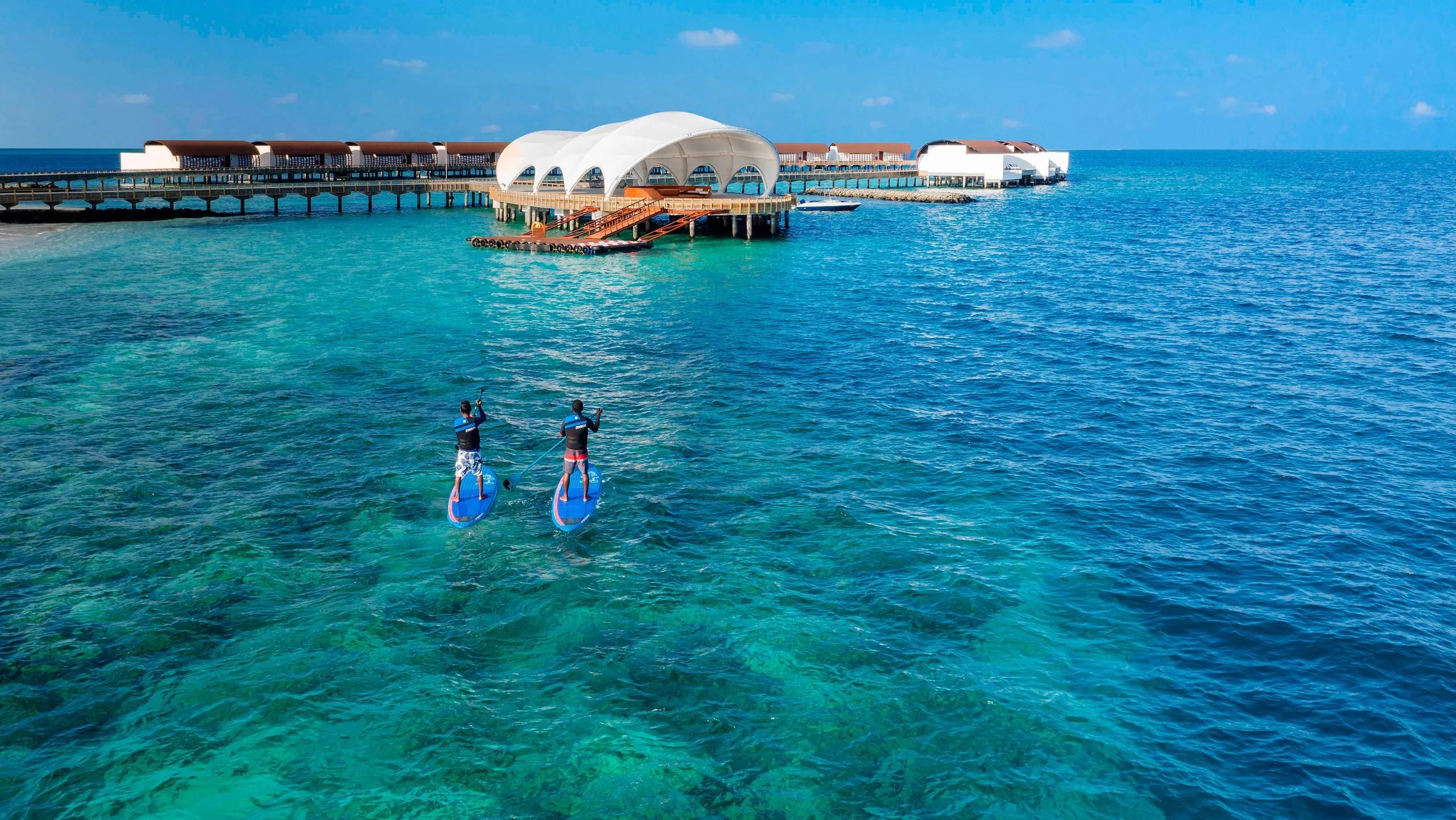 Two guests paddleboarding past the resort's watersports jetty and its white canopy roof