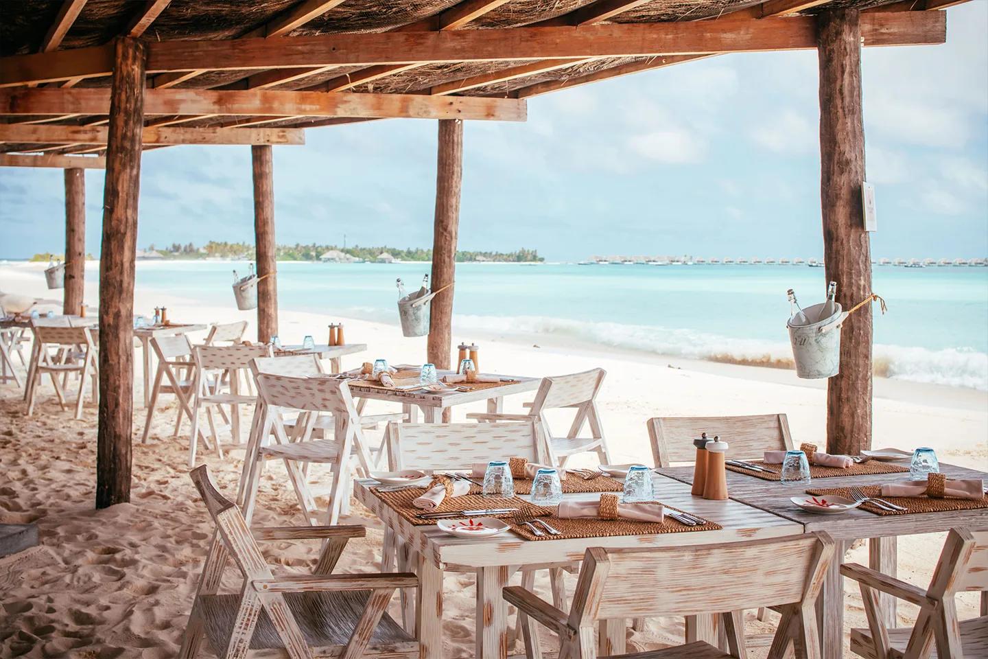 Crab Shack's beachfront dining tables under a rustic timber pergola roof, set directly on the sand with the lagoon and distant overwater villas in view