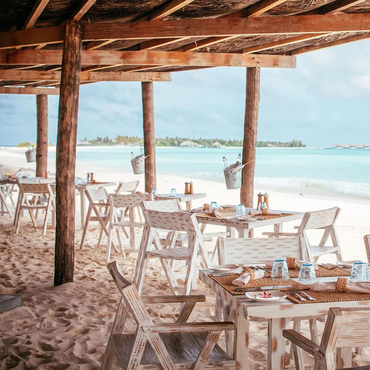 Crab Shack's beachfront dining tables under a rustic timber pergola roof, set directly on the sand with the lagoon and distant overwater villas in view