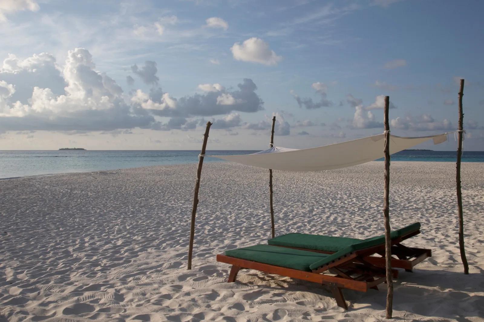 A shaded daybed with green cushions set up on the beach in front of the calm lagoon