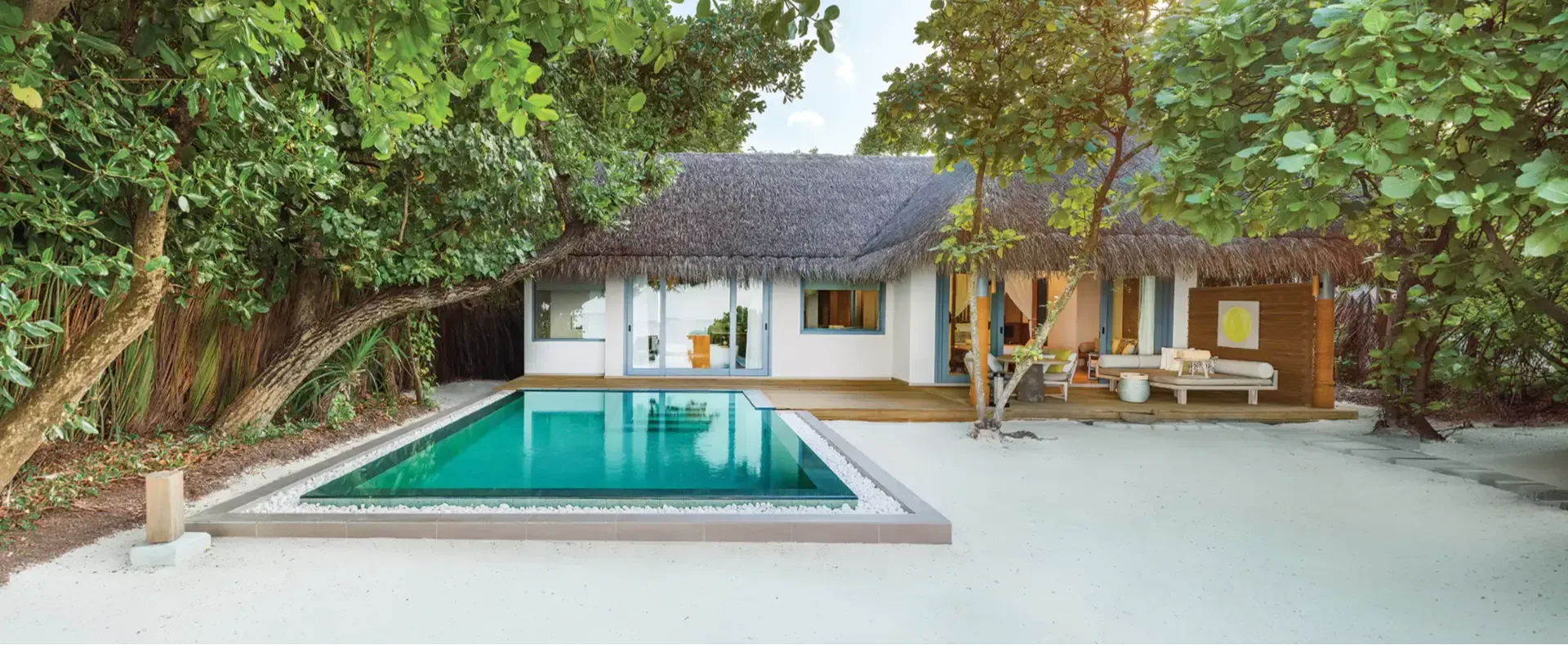 A Beach Pool Villa's rectangular pool in front of a thatched-roof villa, sun deck and lounge area visible through the open doors