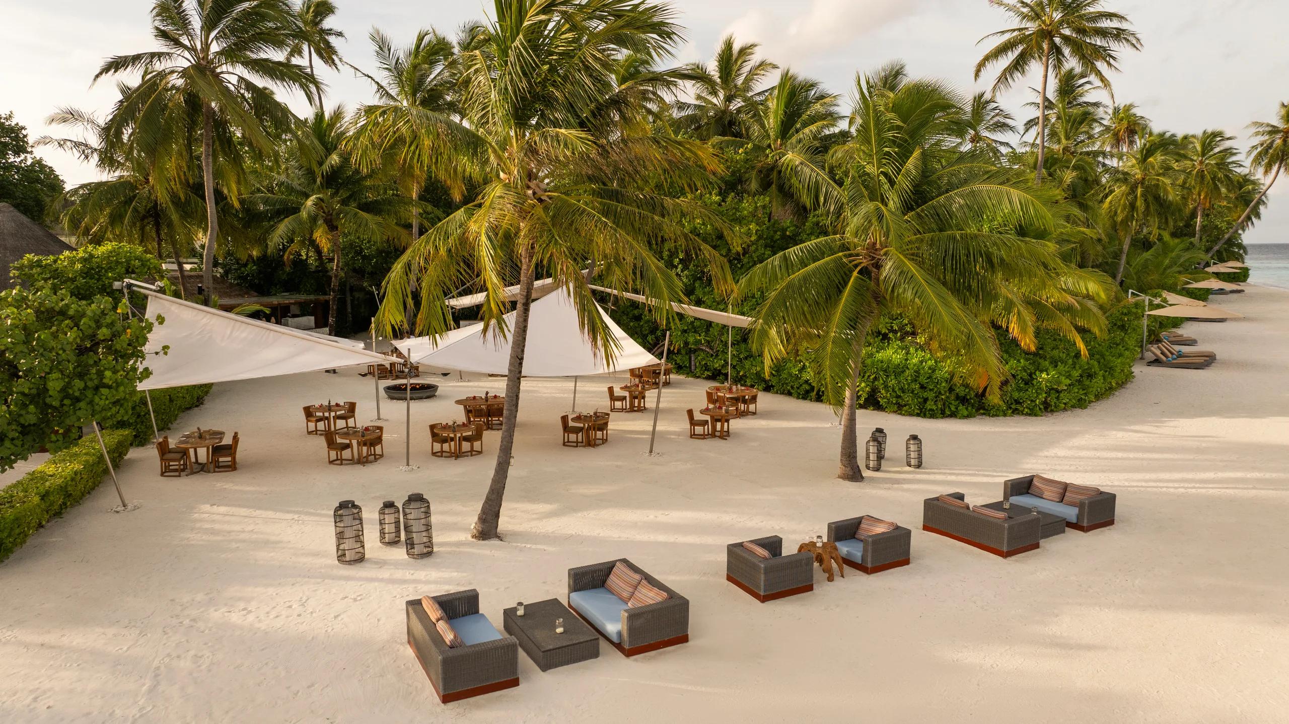 An open-air beachfront terrace shaded by palm trees and white sail canopies, with wooden dining tables set on the sand — Kaika Sushi Bar's outdoor terrace seating