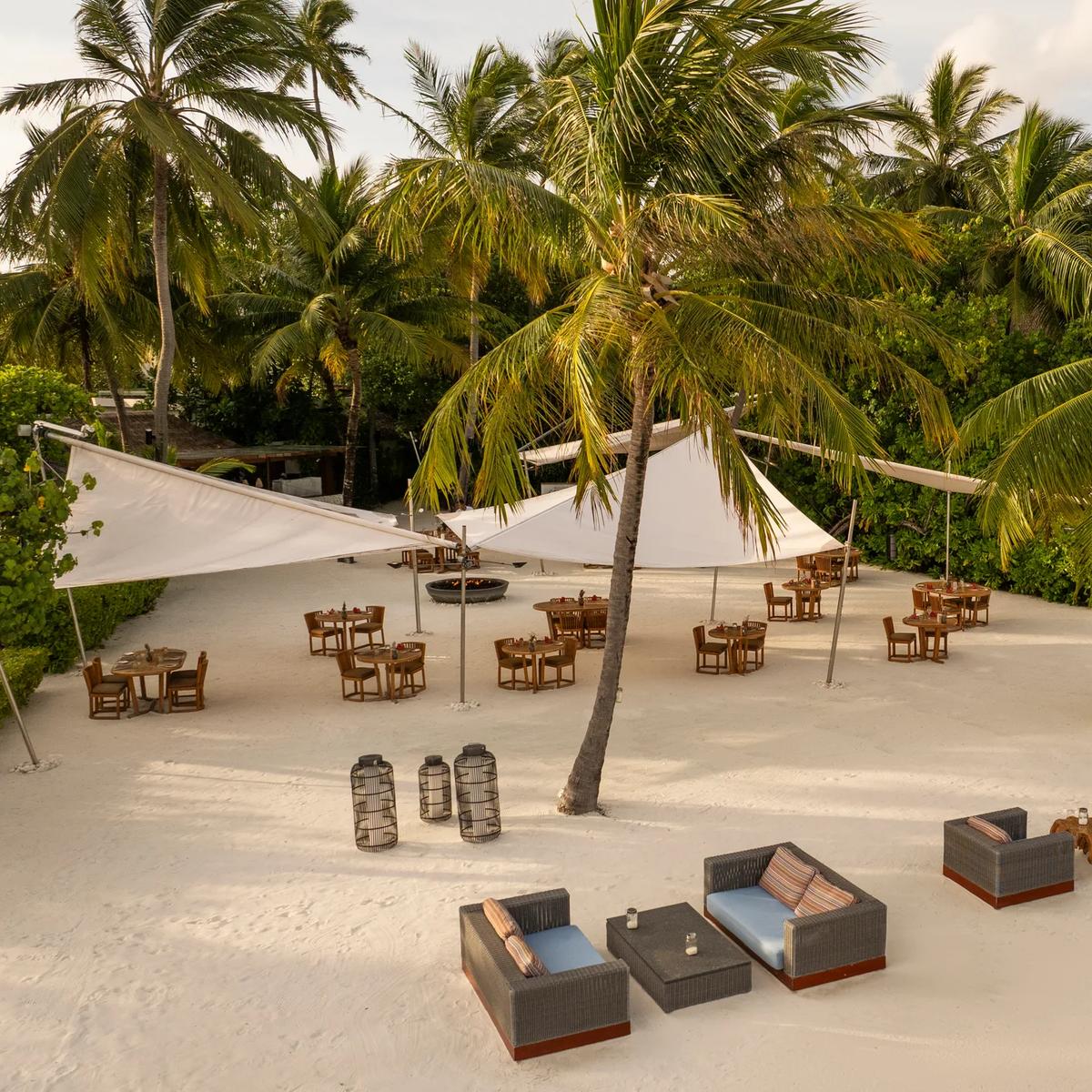 An open-air beachfront terrace shaded by palm trees and white sail canopies, with wooden dining tables set on the sand — Kaika Sushi Bar's outdoor terrace seating