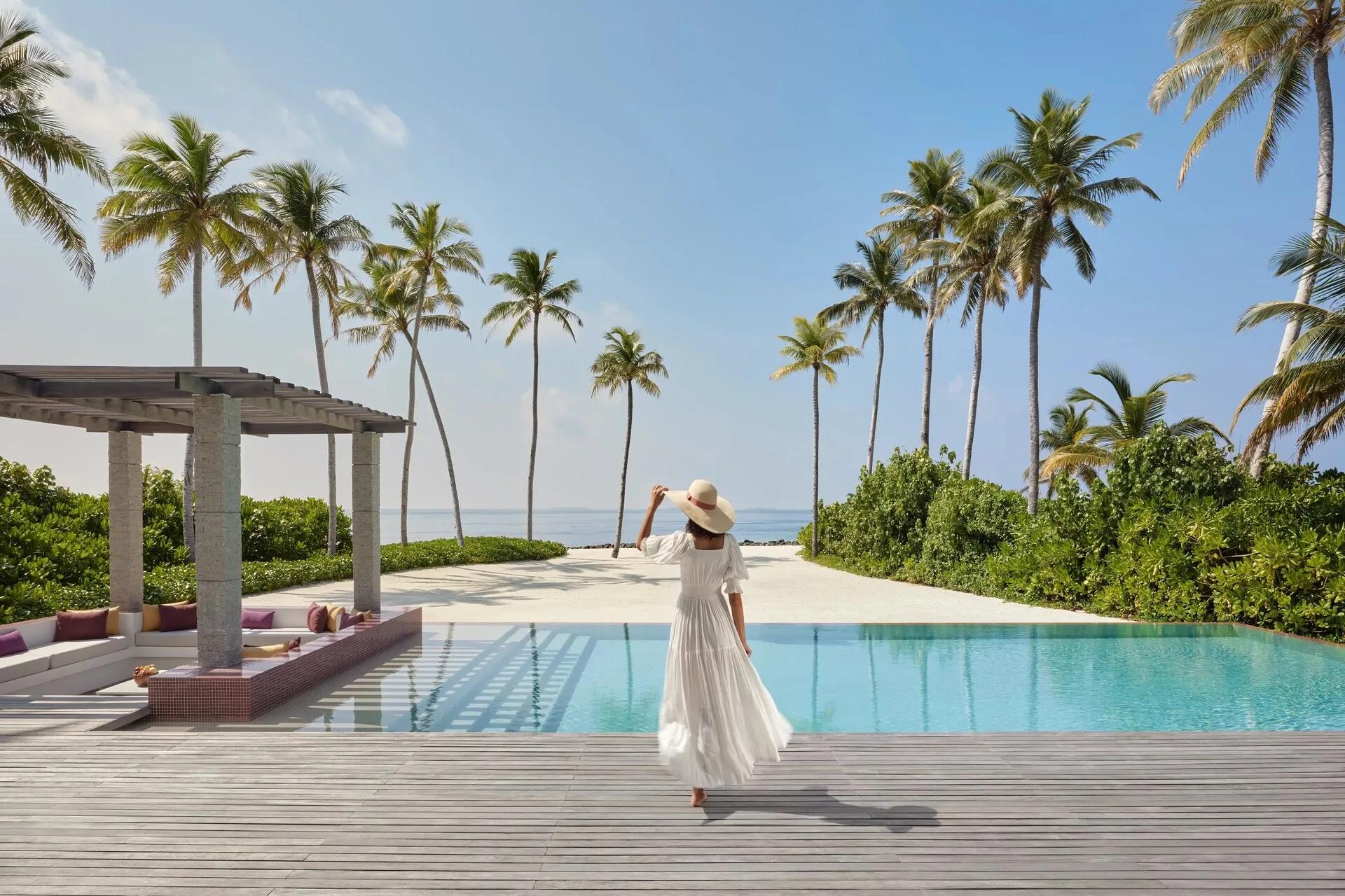 A guest in a wide-brimmed hat walking beside an infinity pool lined with palm trees, the lagoon visible beyond