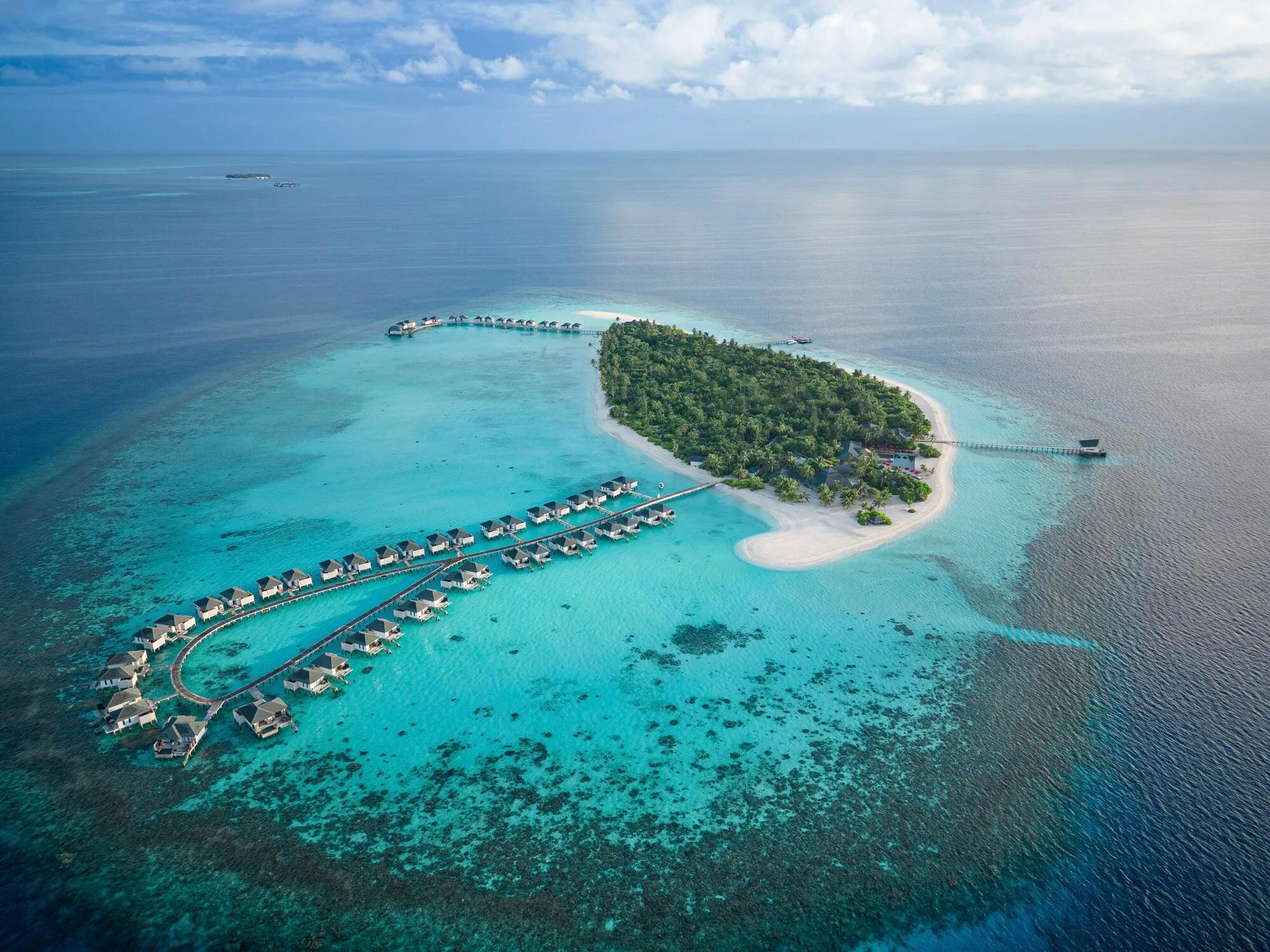 Aerial view of Havodda's point, a wooden jetty reaching from an overwater villa into the reef, with a thatched pool deck, red umbrellas and loungers set into the beach below