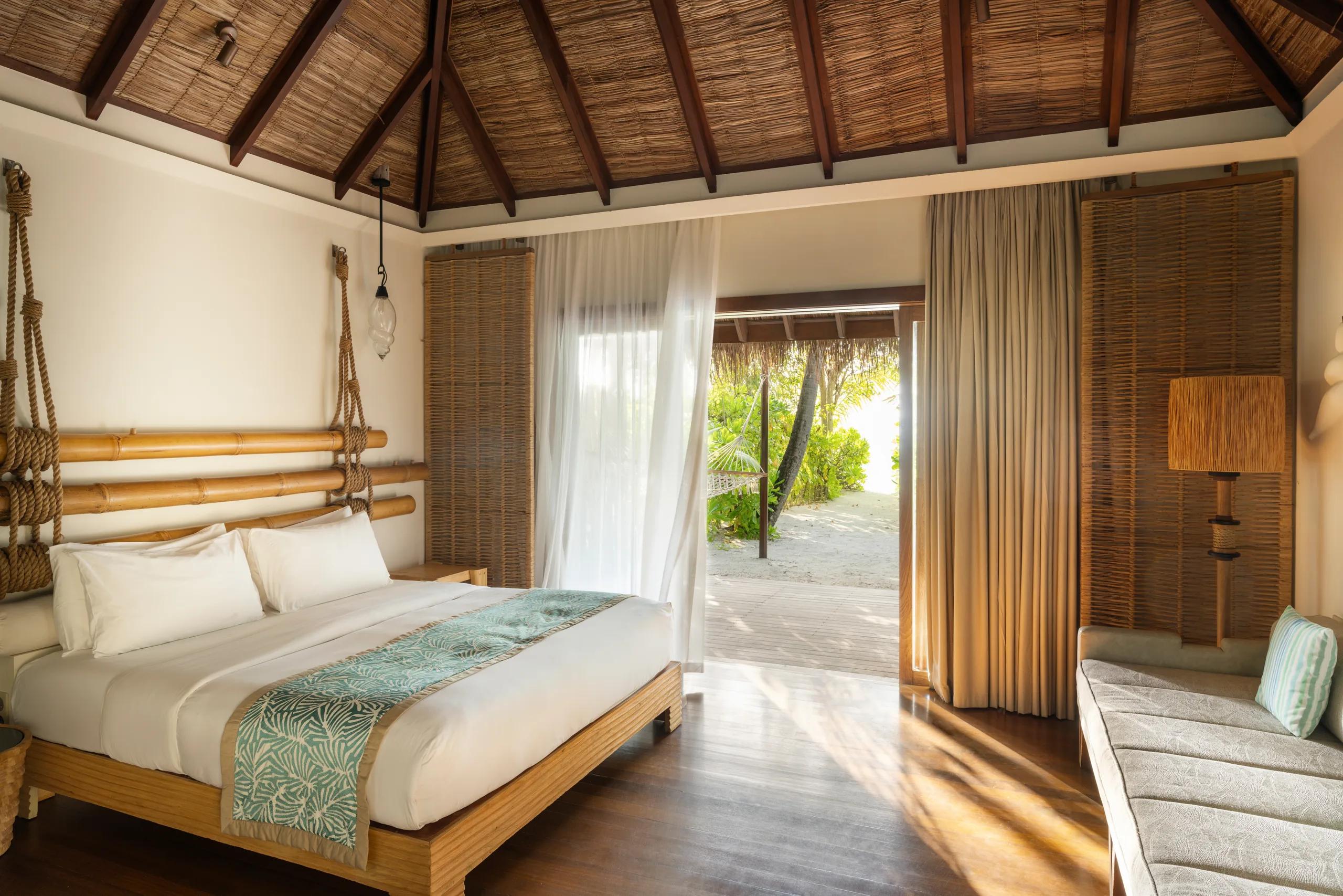 A Beach Villa bedroom with a rope-and-bamboo headboard, sheer curtains drawn back to a garden deck with a hammock and sunloungers