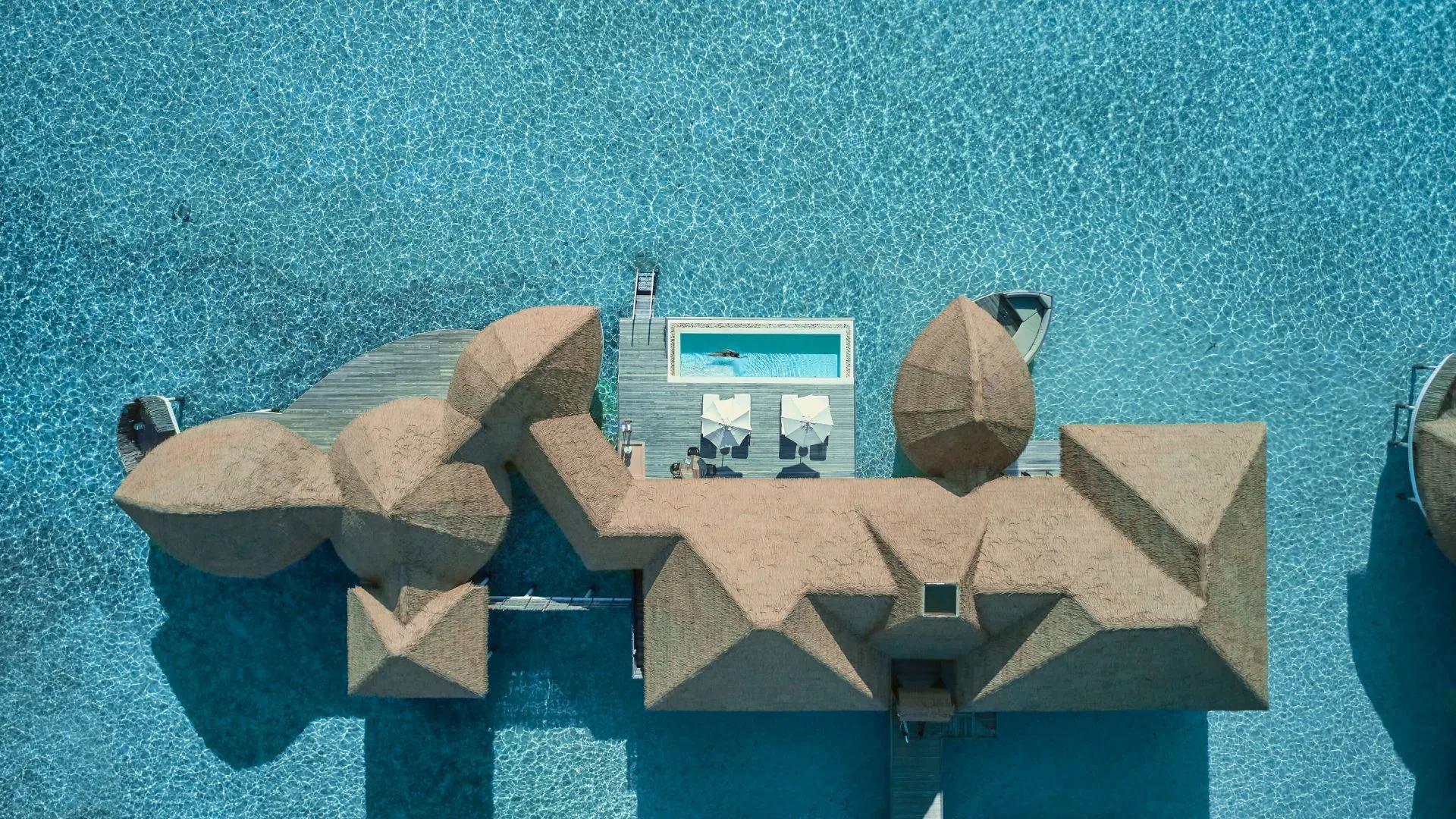 Directly overhead view of an overwater villa's thatched roof and private plunge pool, with a swimmer in the pool and a boat moored alongside