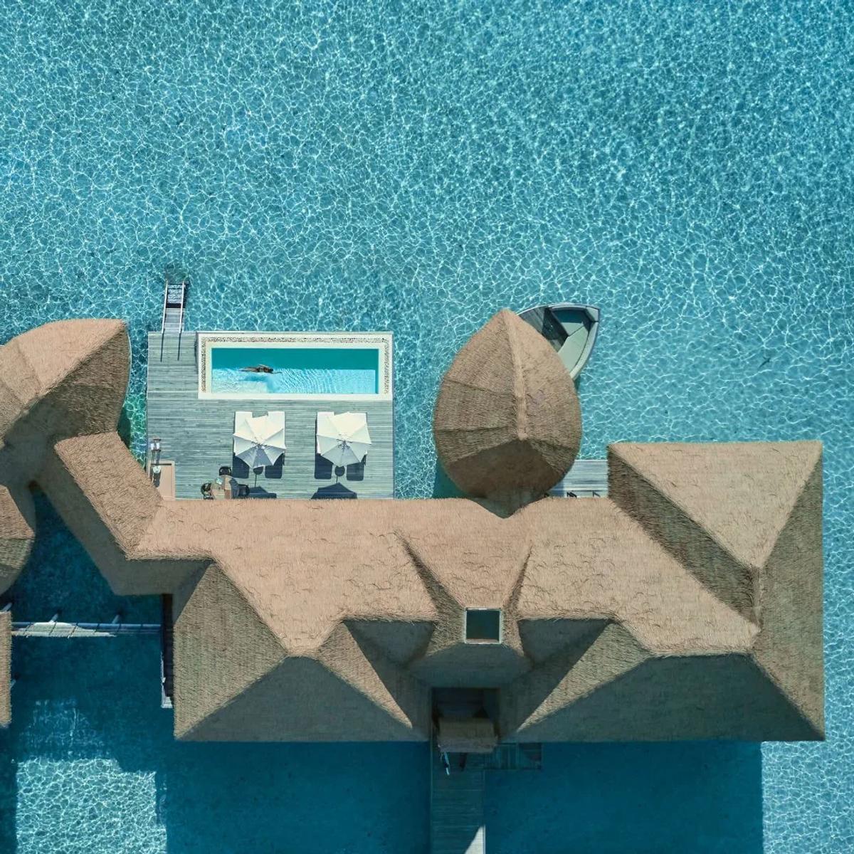 Directly overhead view of an overwater villa's thatched roof and private plunge pool, with a swimmer in the pool and a boat moored alongside