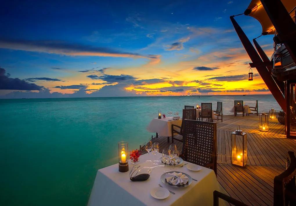 Tables set for dinner on The Lighthouse Restaurant's overwater deck at dusk, lanterns lit as the sky turns orange over the lagoon