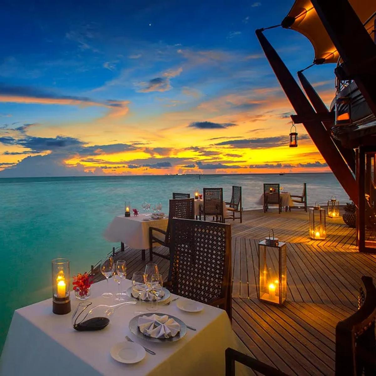 Tables set for dinner on The Lighthouse Restaurant's overwater deck at dusk, lanterns lit as the sky turns orange over the lagoon