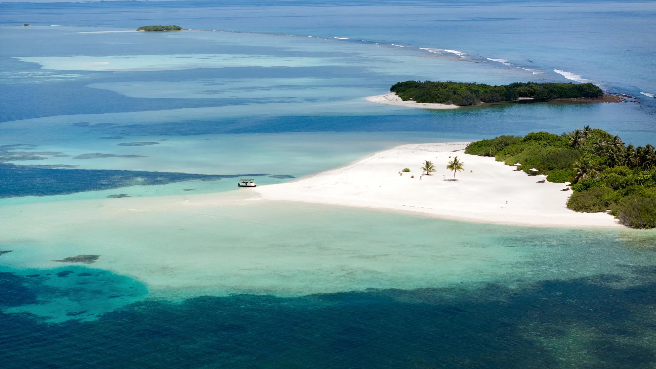 Aerial view of a small sandy islet with a boat pulled up and a few guests on the beach, the arrival point for a castaway lunch.