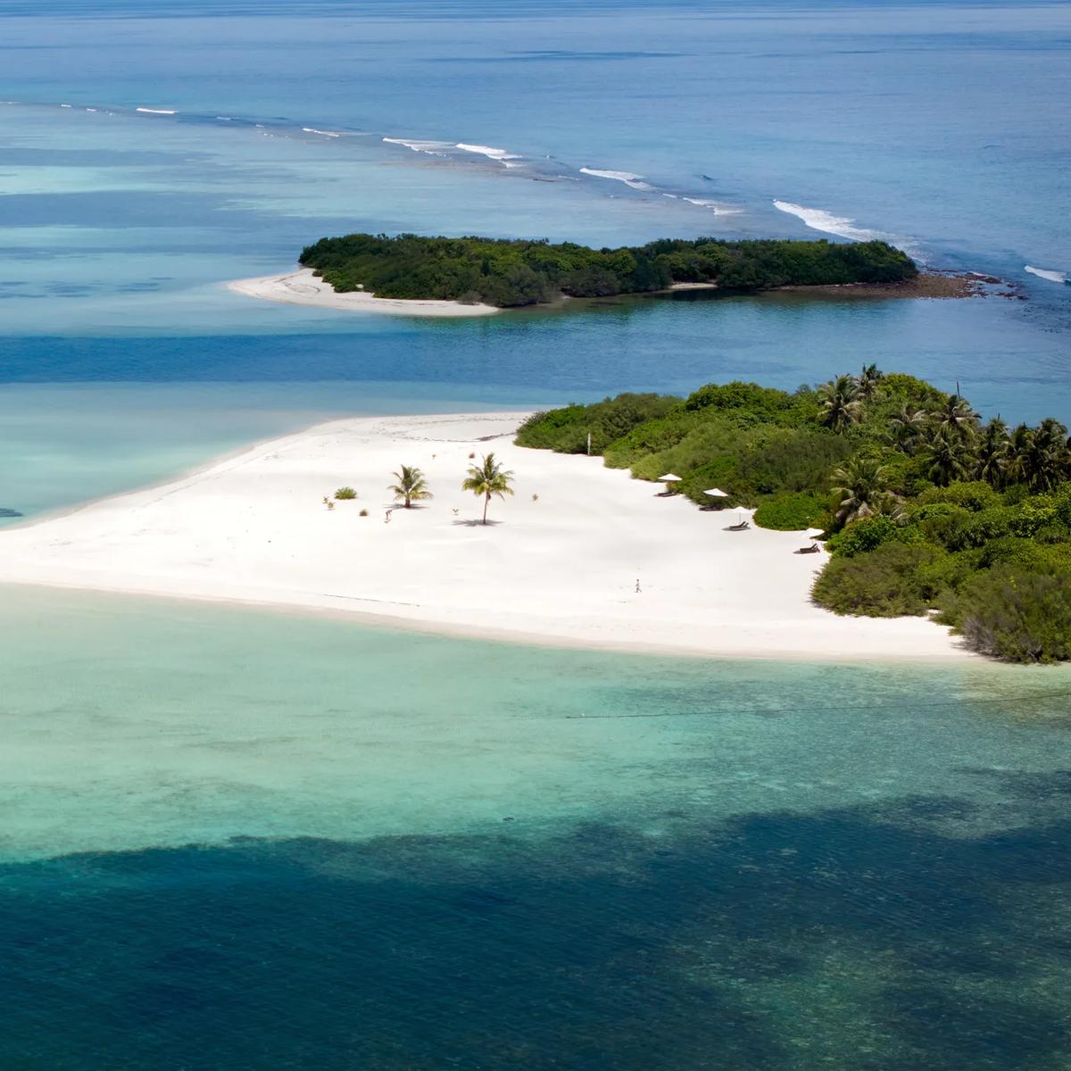 Aerial view of a small sandy islet with a boat pulled up and a few guests on the beach, the arrival point for a castaway lunch.