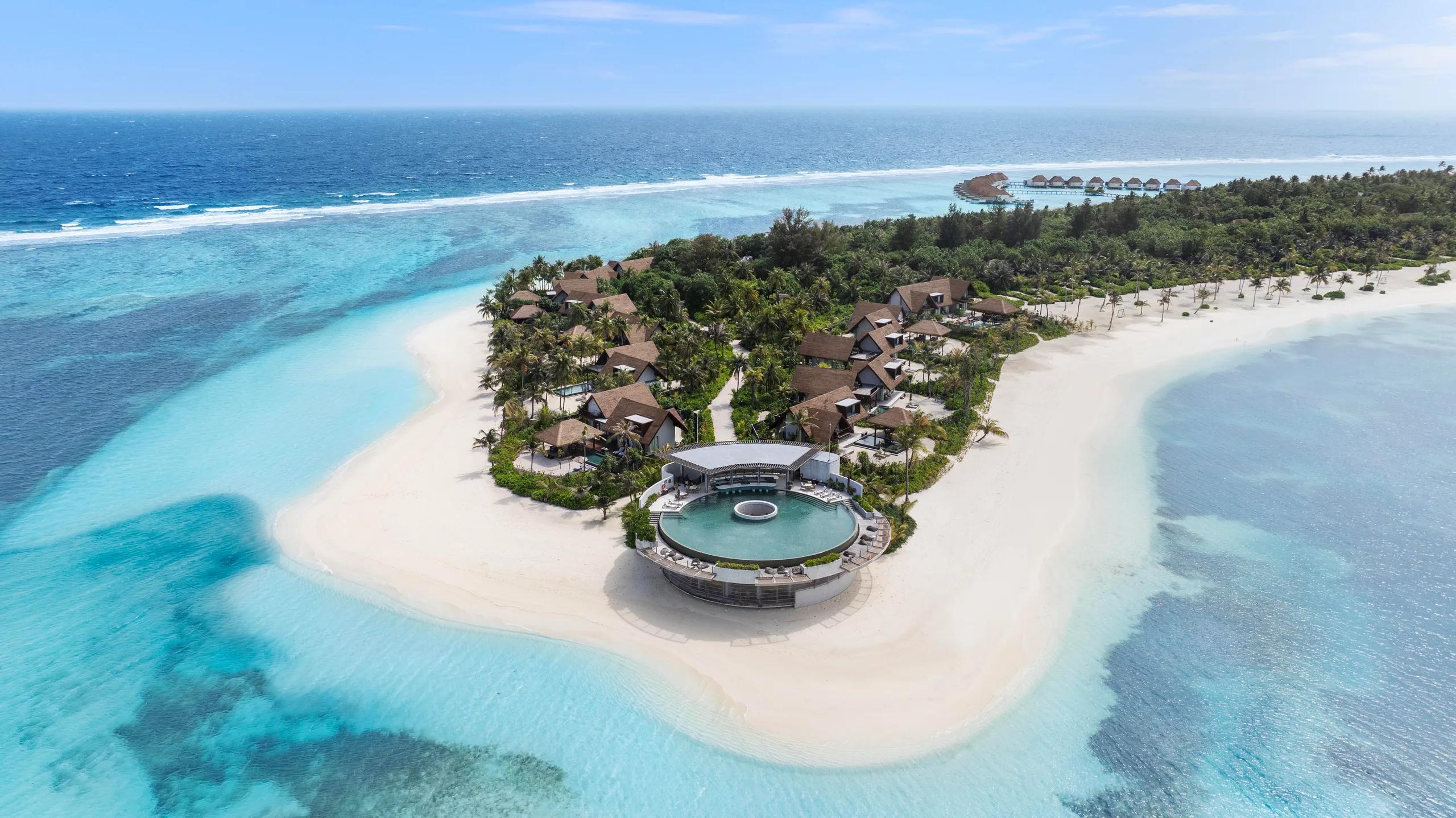 Aerial view of the round infinity pool and dining pavilion at the point of Kanuhura island, framed by overwater villas and reef.