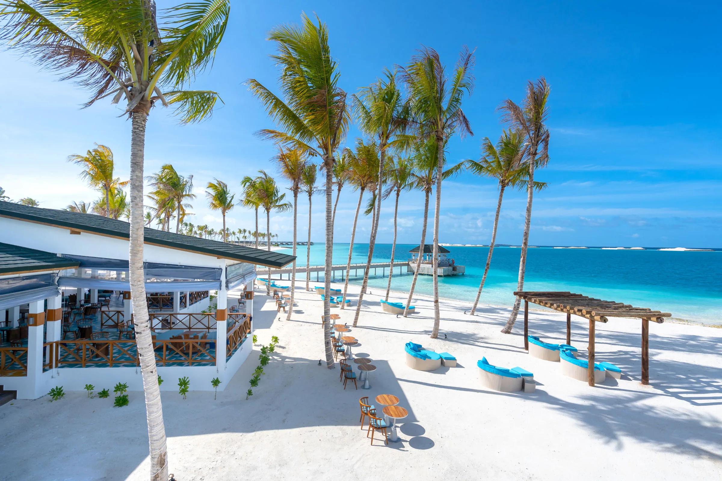 Blue daybeds and dining tables along Rainbow's beachfront terrace, a jetty reaching out to an overwater pavilion beyond