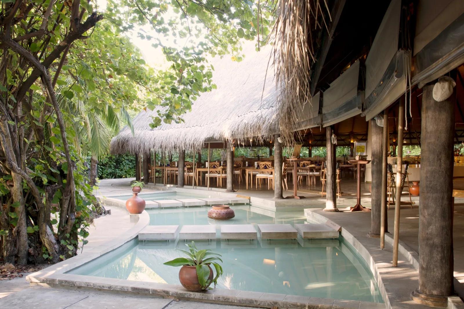 Cowrie restaurant's open-air dining pavilion, reflecting pool along the walkway and tables set beneath a high thatched roof