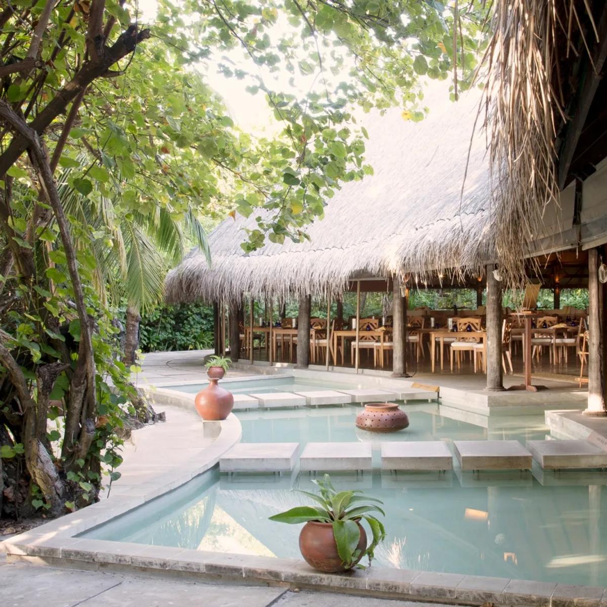 Cowrie restaurant's open-air dining pavilion, reflecting pool along the walkway and tables set beneath a high thatched roof