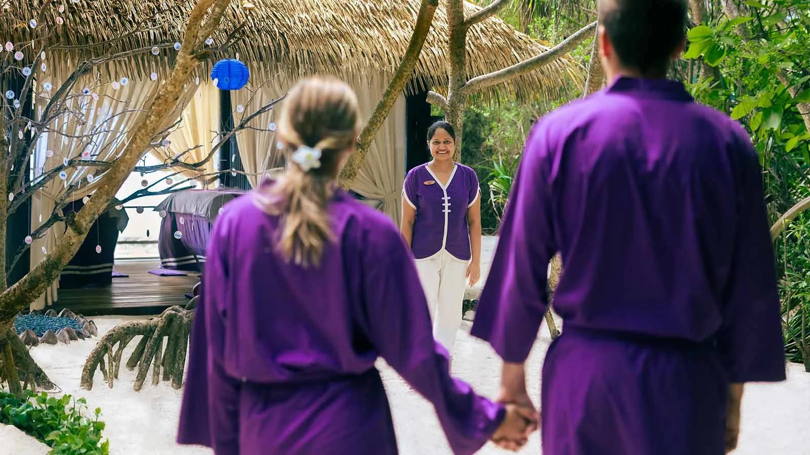 Two guests in purple robes walking hand in hand towards a thatched treatment pavilion at Duniye Spa