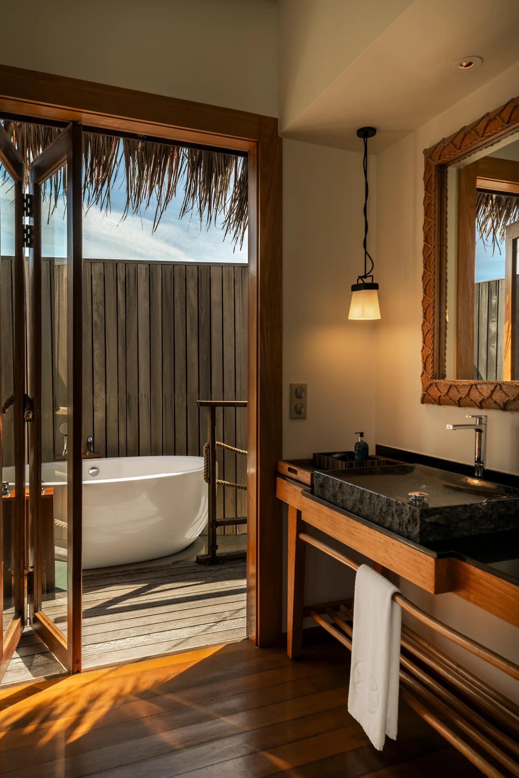 A free-standing white outdoor bathtub on a wooden villa deck, screened by timber slat walls