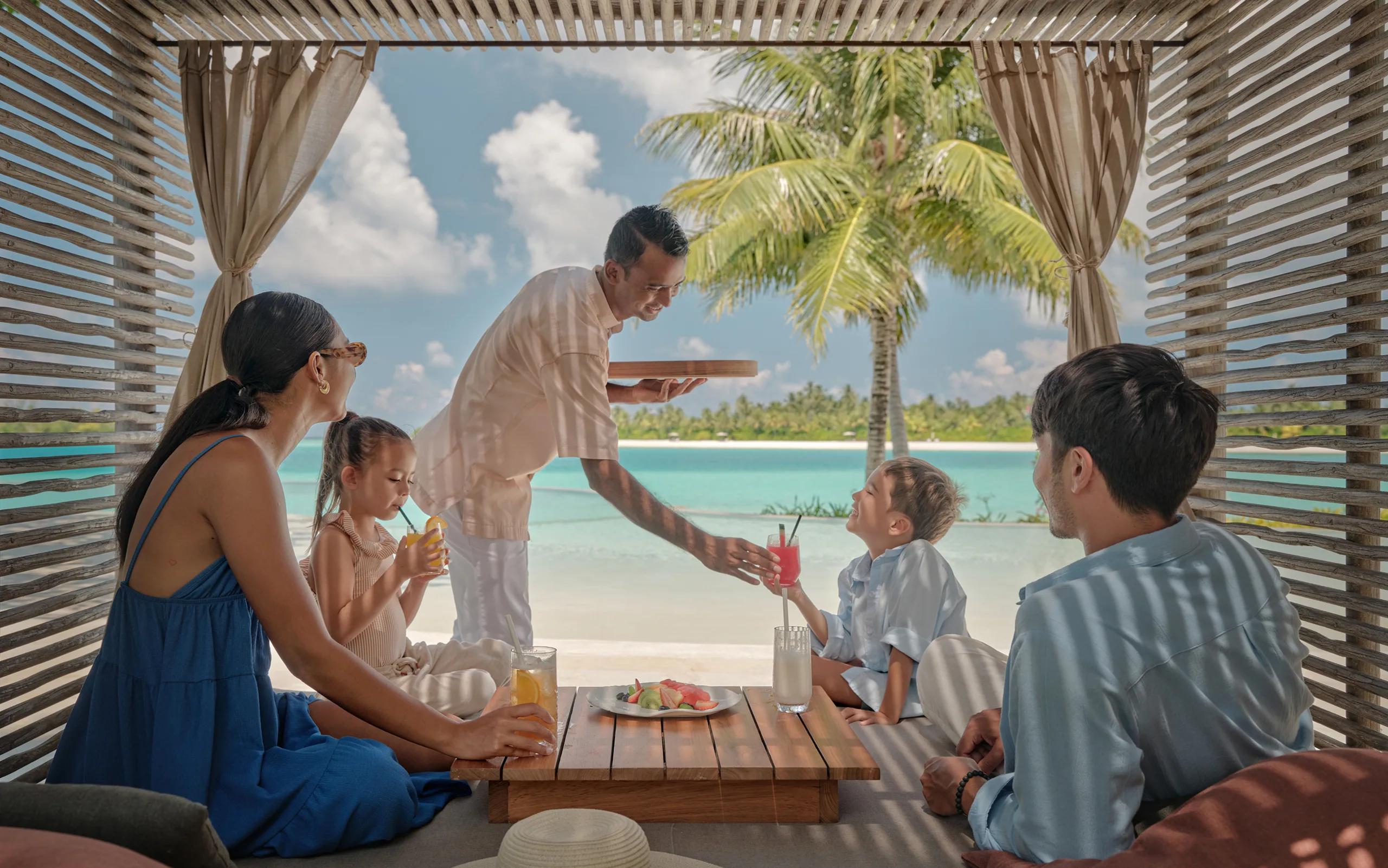 A waiter serves drinks to a family relaxing in a shaded cabana at Fari Beach Club, the lagoon and a palm tree beyond.