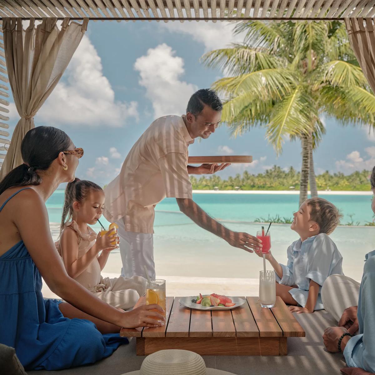 A waiter serves drinks to a family relaxing in a shaded cabana at Fari Beach Club, the lagoon and a palm tree beyond.
