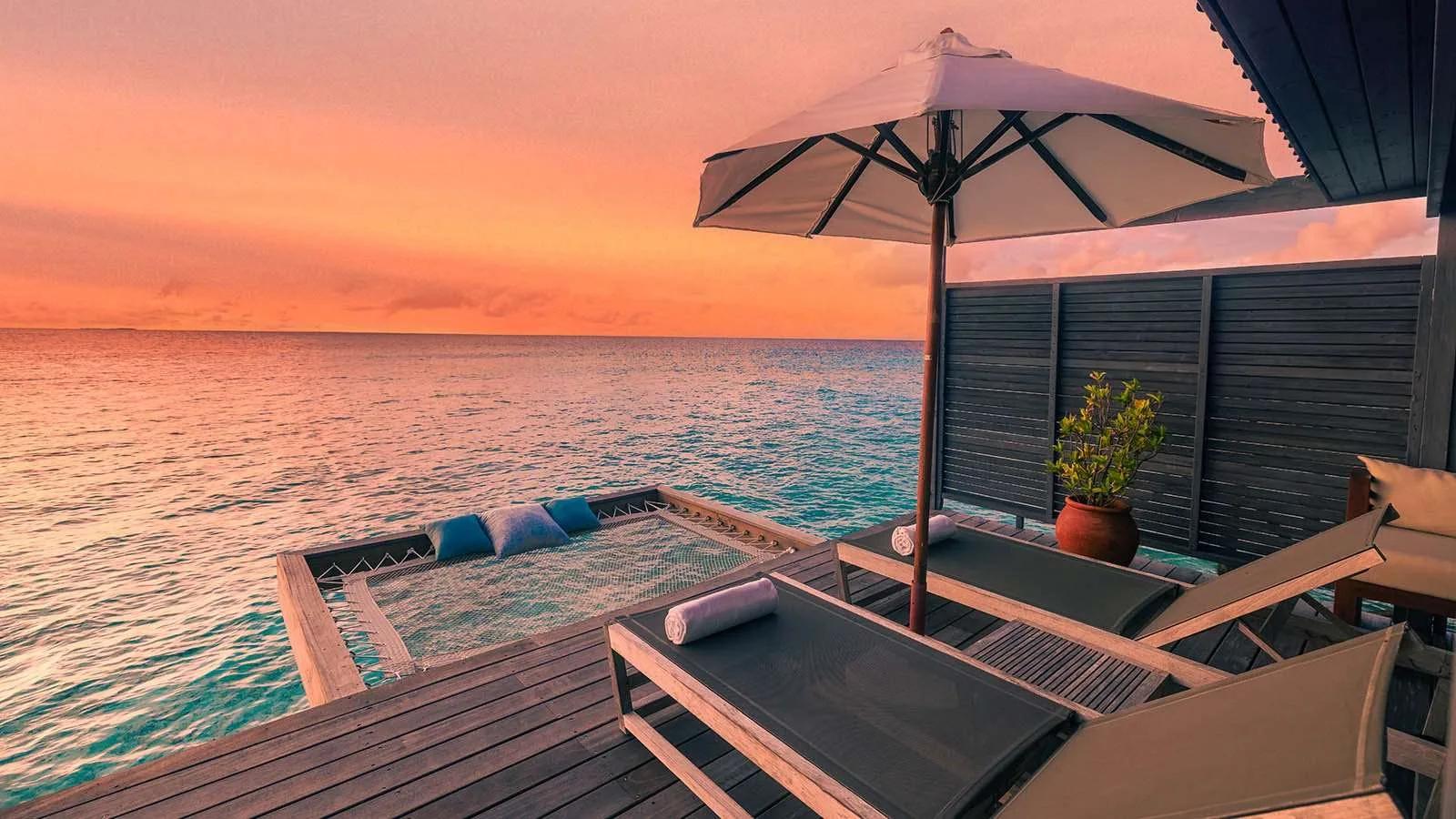 The deck of an Ocean Jacuzzi Water Villa at sunset, a catamaran-net hammock and lounge chairs set over the water