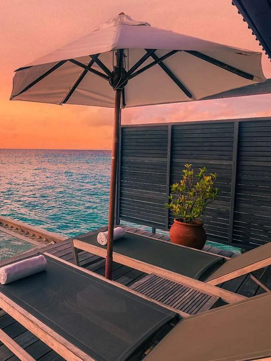 The deck of an Ocean Jacuzzi Water Villa at sunset, a catamaran-net hammock and lounge chairs set over the water