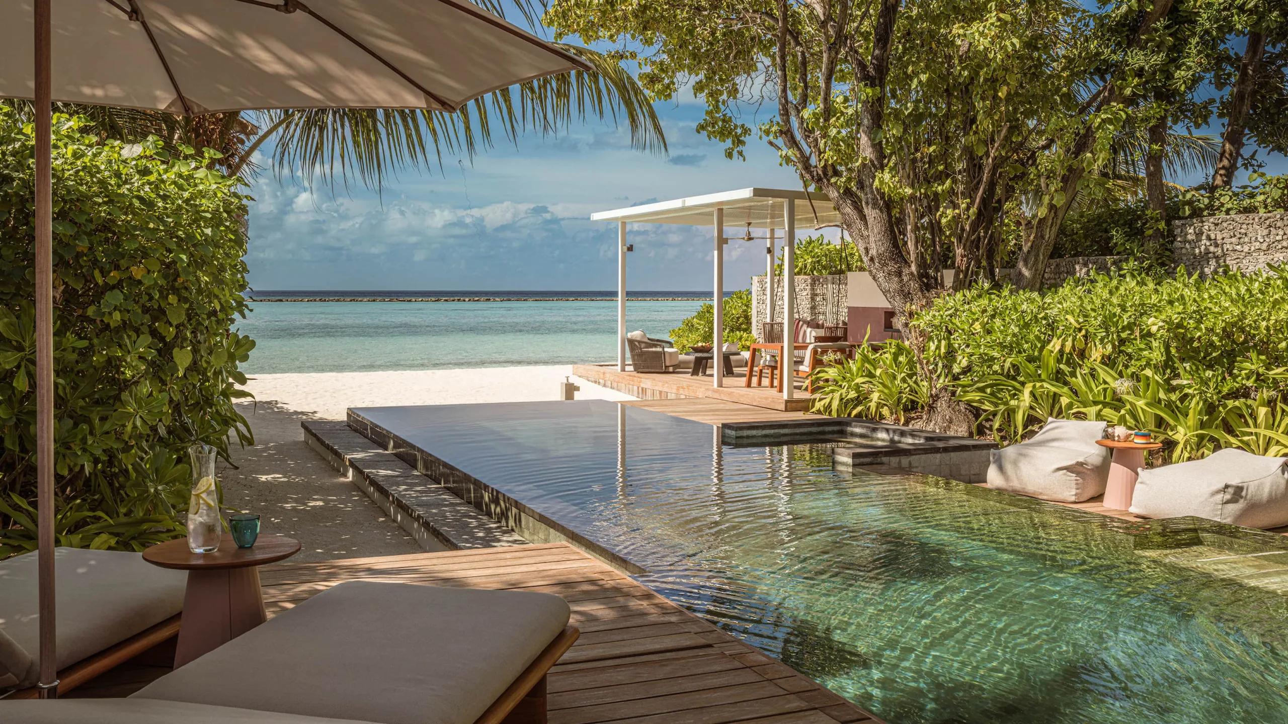 A Family Oceanfront Bungalow's pool deck beneath palm trees, with a shaded dining pavilion and beach visible through the foliage
