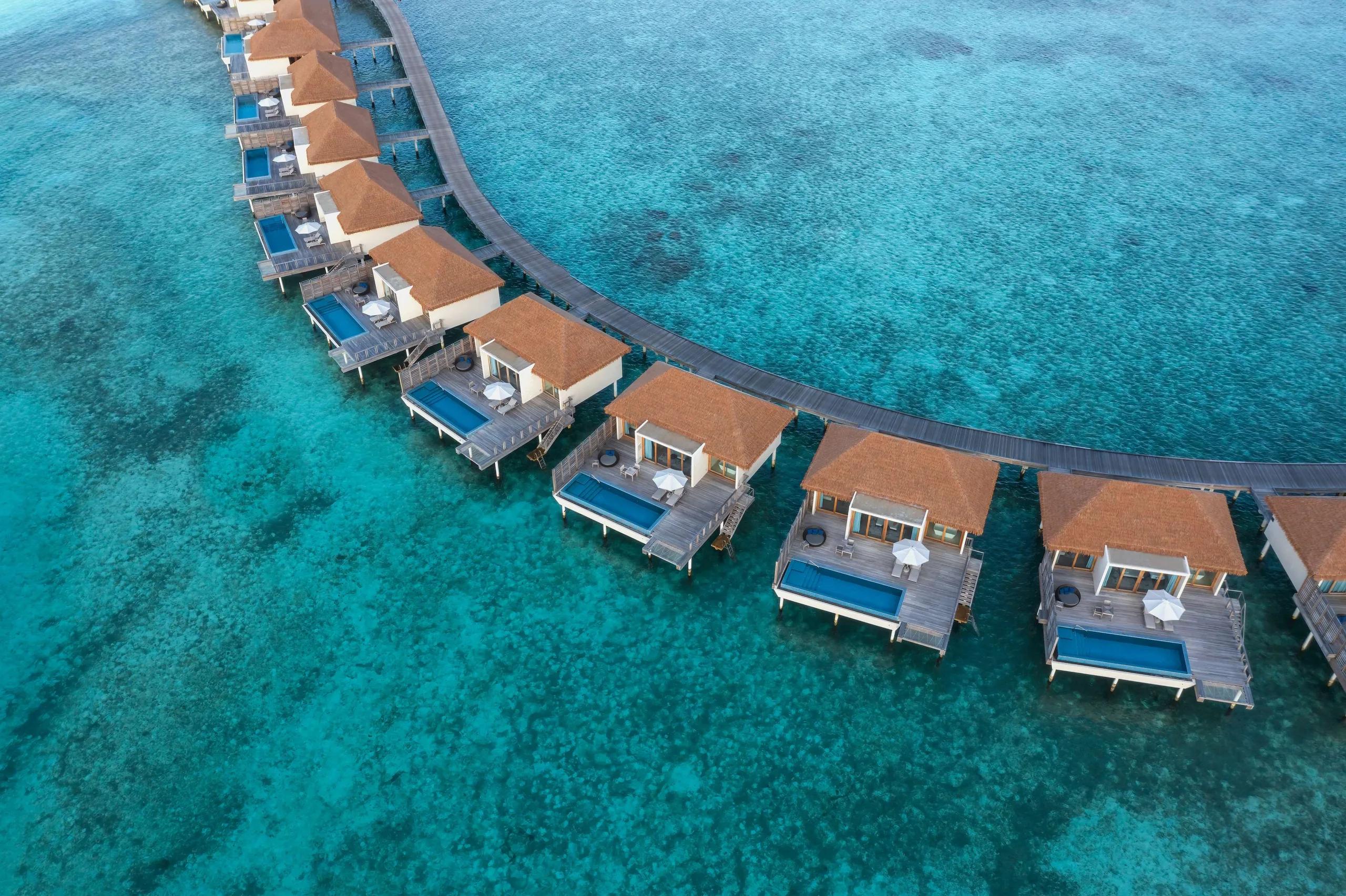 Radisson Blu Resort Maldives' curved row of overwater villas seen directly from above, private pools and a wooden jetty tracing the reef edge