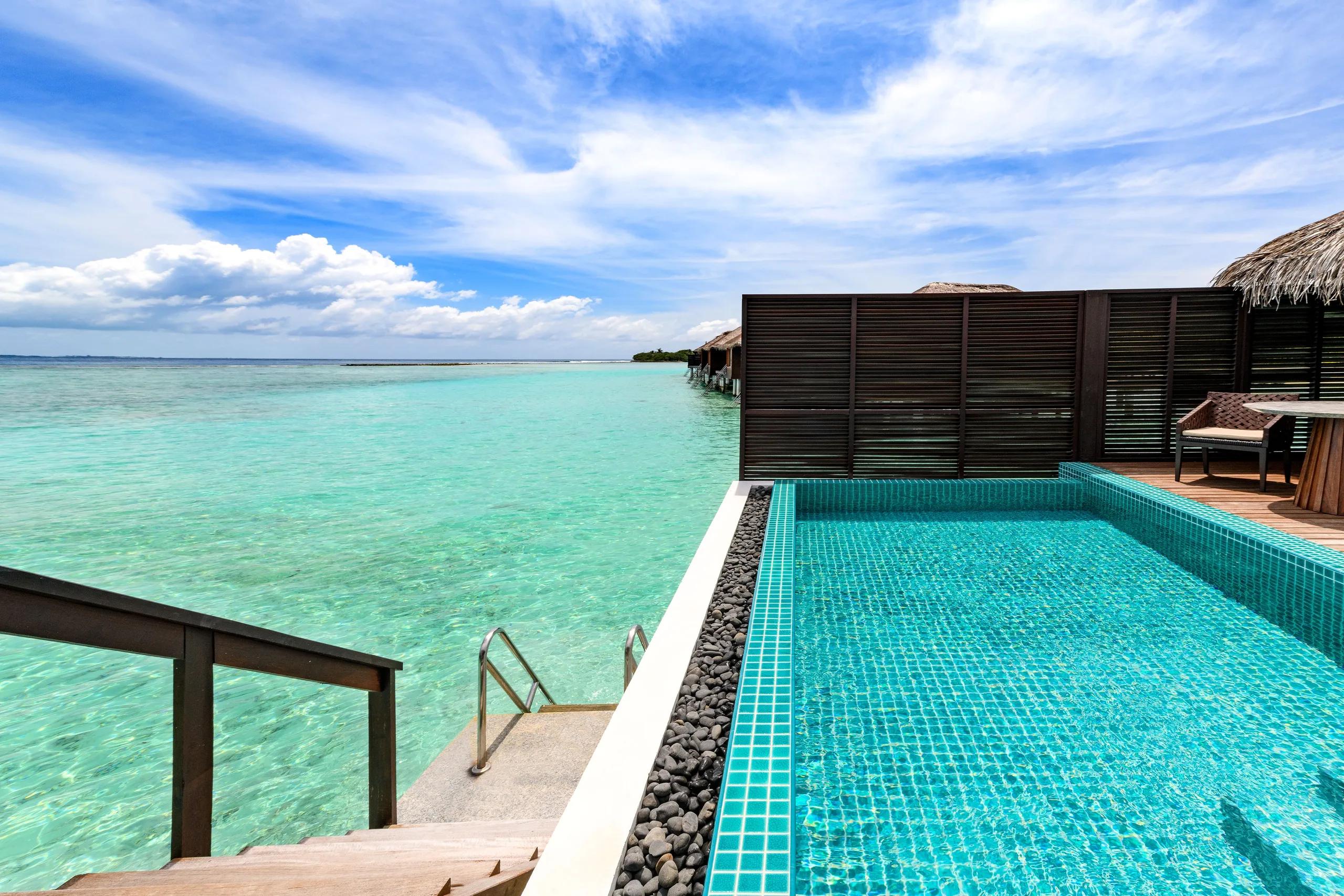 A long wooden jetty leading from the beach to a row of thatched-roof overwater villas, seen from ground level