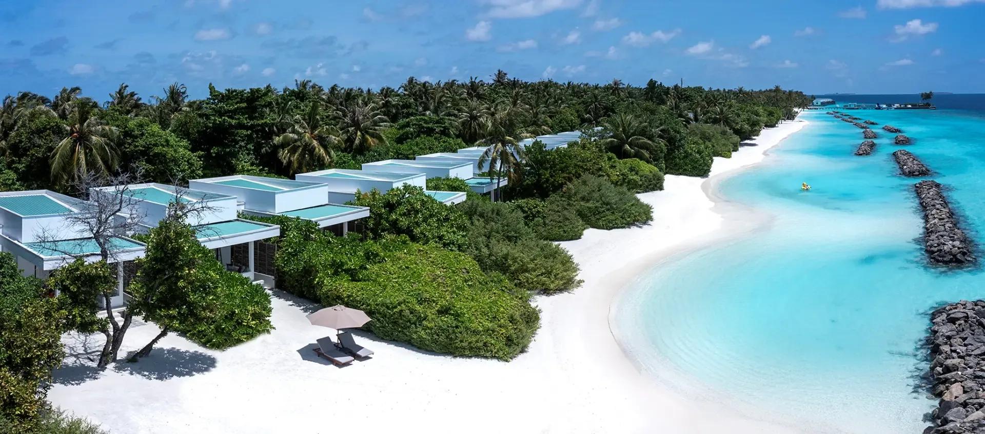 Panoramic aerial of Niva Dhigali's beachfront villa row curving toward the overwater villas and reef