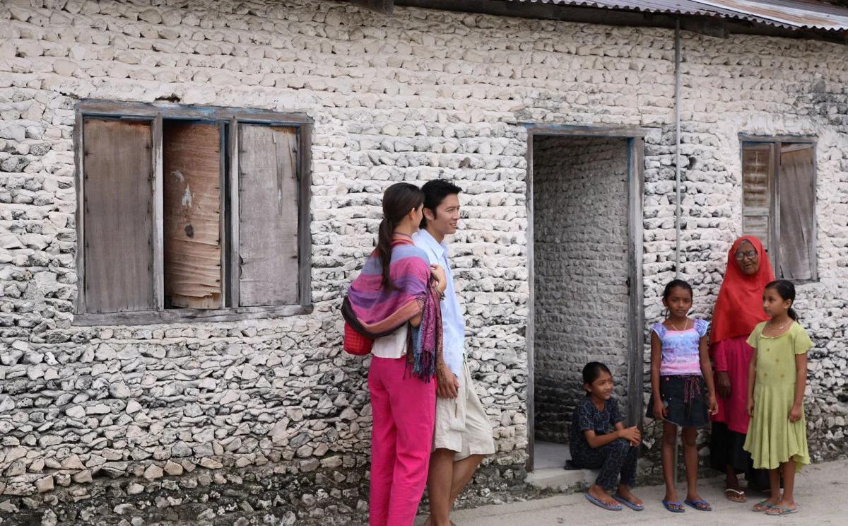 A couple greeted by a local family outside a coral-stone house on a nearby inhabited island