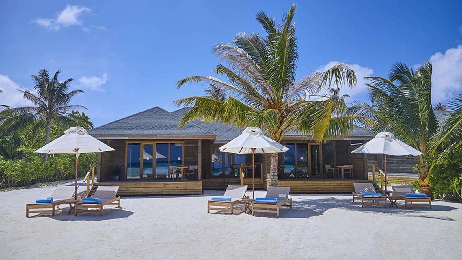 The exterior of a Dheru Island Three Bedroom Villa, three loungers and umbrellas set along its beachfront deck