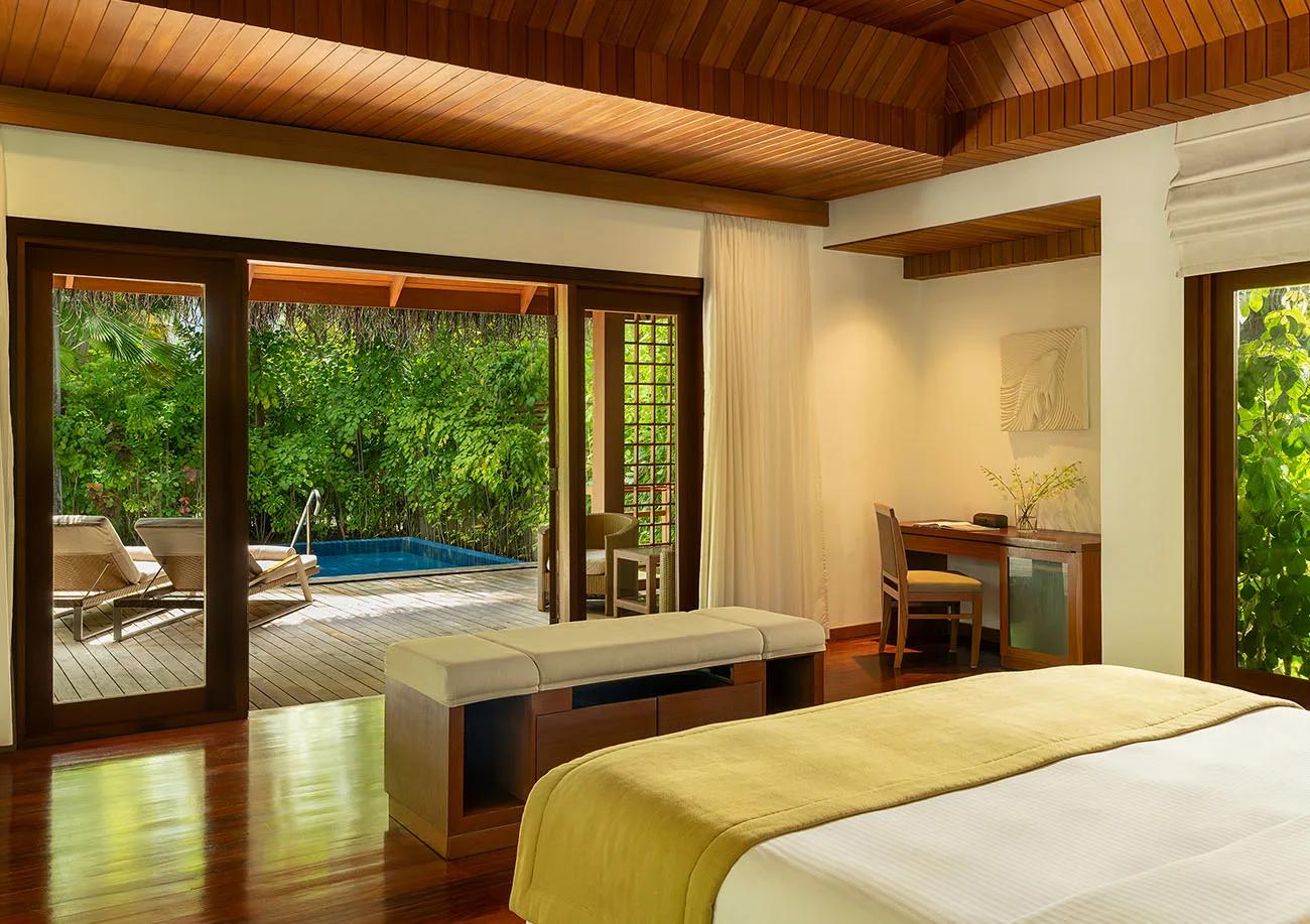 A Baros Pool Villa bedroom opening onto its private pool deck and sun loungers, screened by tropical garden foliage