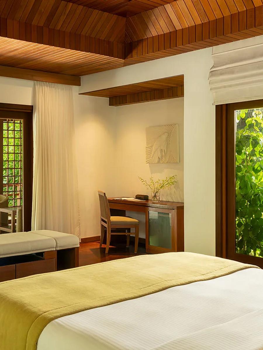 A Baros Pool Villa bedroom opening onto its private pool deck and sun loungers, screened by tropical garden foliage