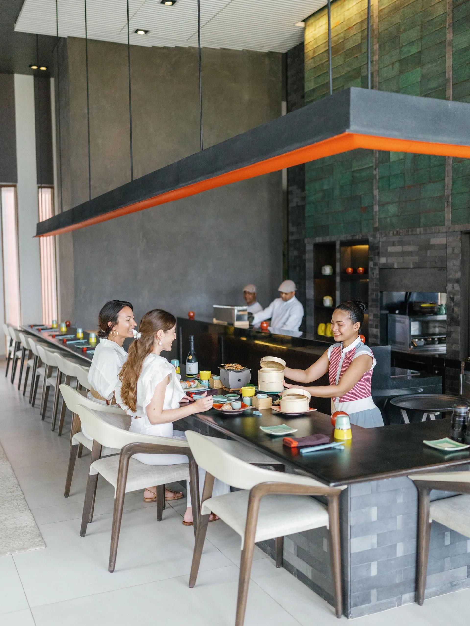 Chefs at The Diptyque's live-cooking counter serving bamboo steamers to a couple seated at the bar