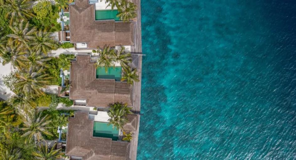 Aerial view of thatched-roof houses and private pools lined along the shoreline, reef visible in the turquoise water beyond