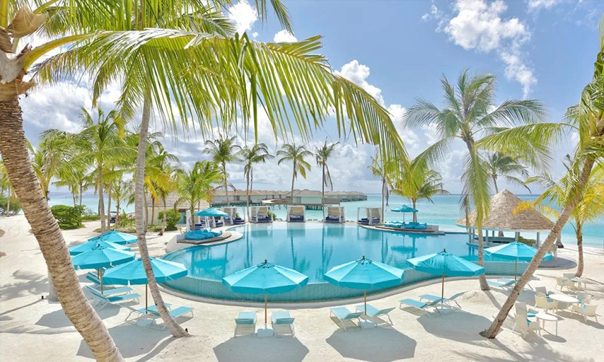Kandima's main pool framed by palms, with the overwater villa jetty visible in the lagoon beyond