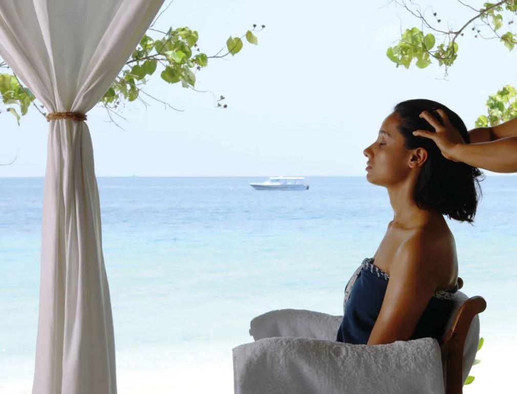 Woman receiving a head massage in an open-air spa pavilion overlooking the ocean, a boat visible offshore