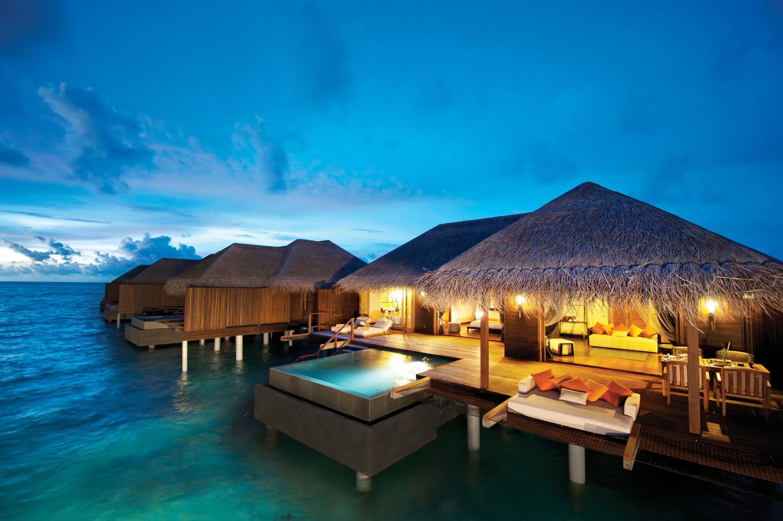 A row of Sunset Ocean Suites lit at dusk, overwater decks and plunge pools above the lagoon