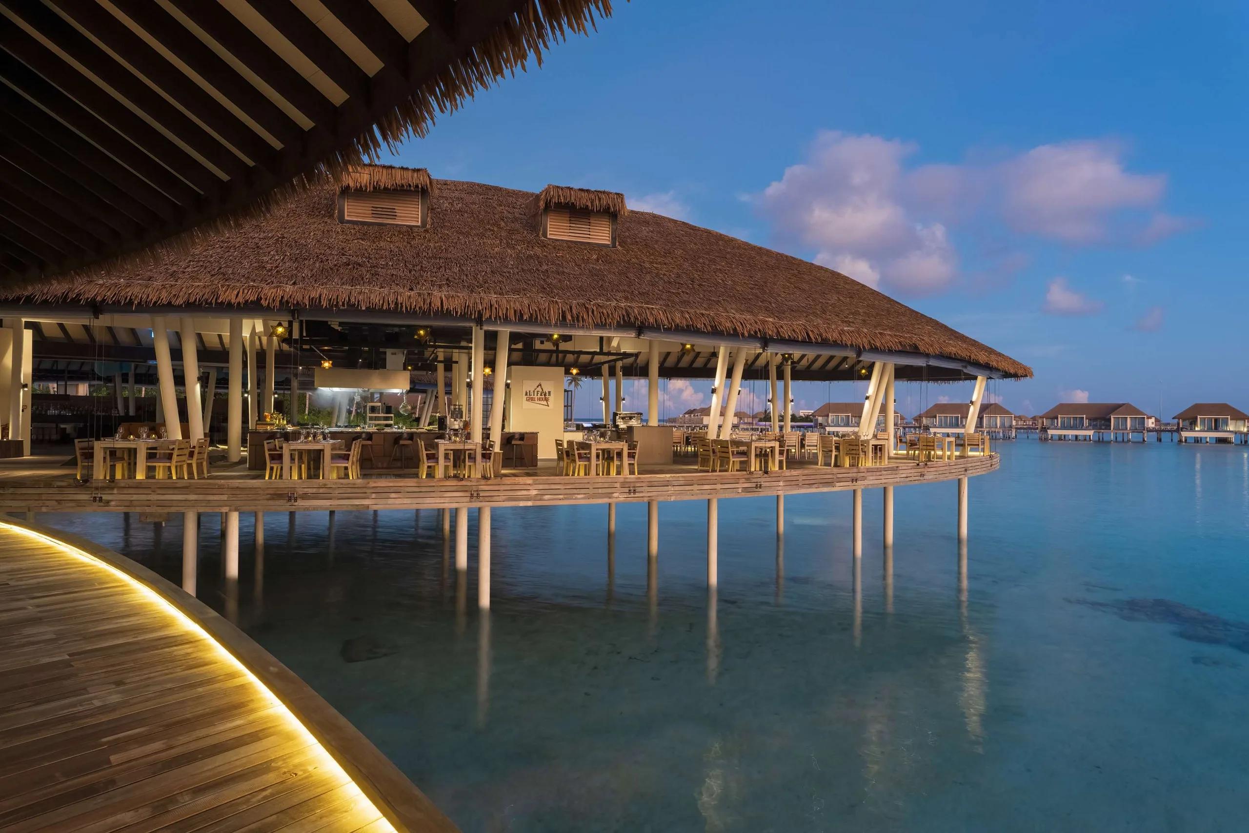 Alifaan's overwater grill house at Radisson Blu Resort Maldives at dusk, its thatched roof lit from within and tables set along the deck