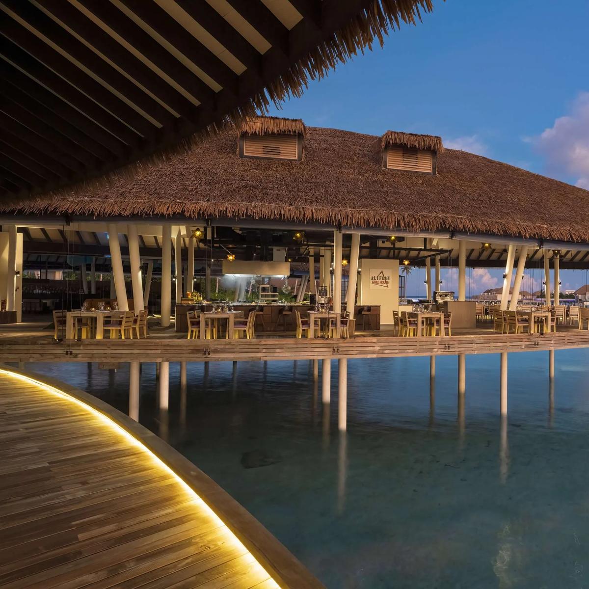 Alifaan's overwater grill house at Radisson Blu Resort Maldives at dusk, its thatched roof lit from within and tables set along the deck