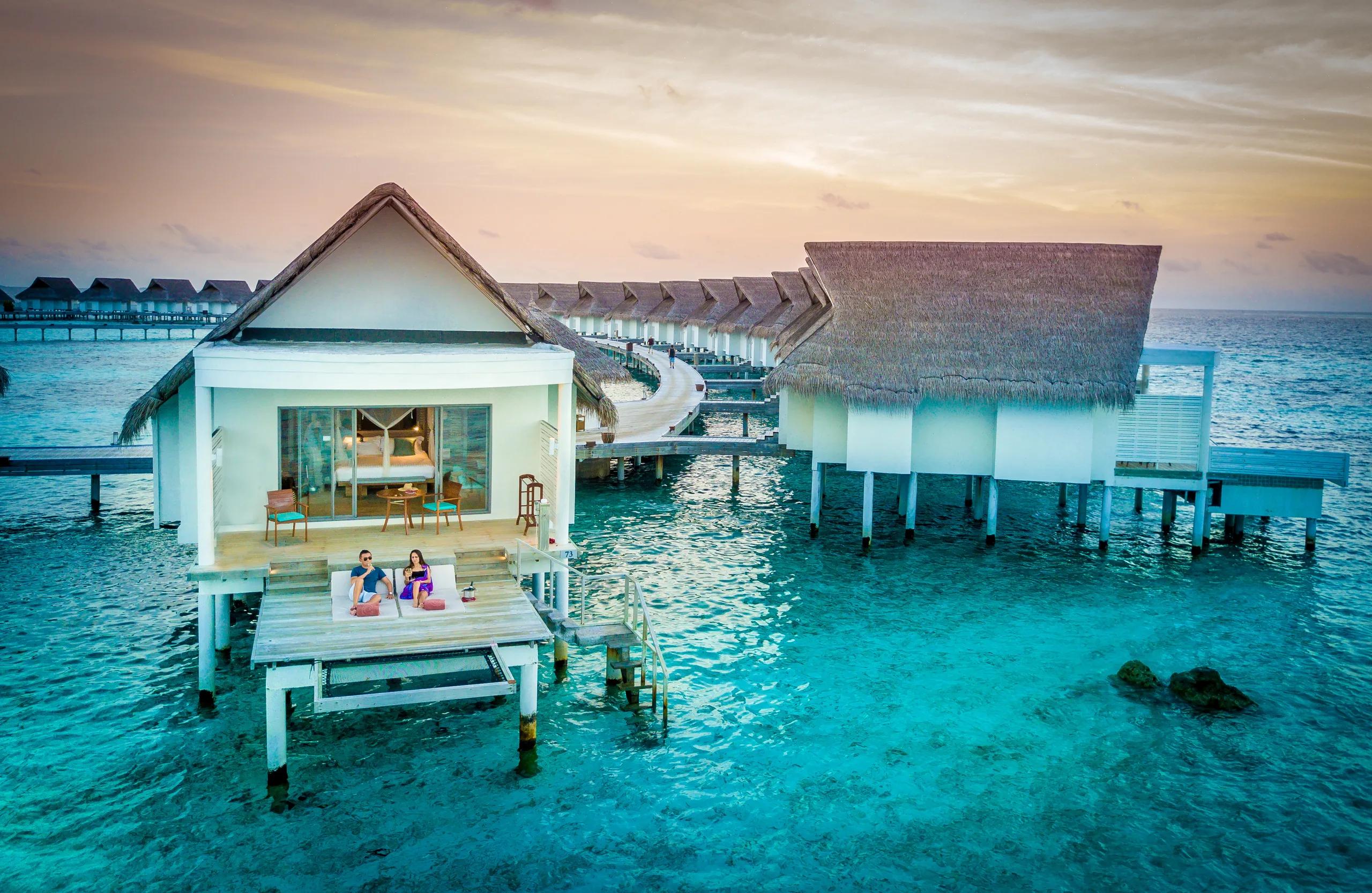 Overwater villas curving out from the island at dusk, a couple relaxing on a private deck in the foreground