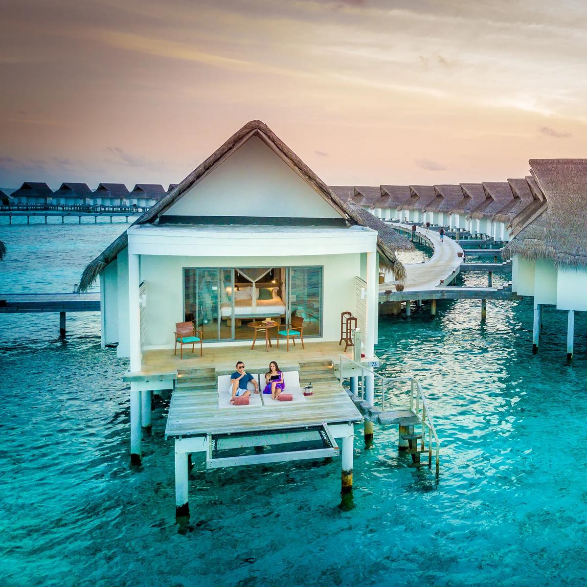 Overwater villas curving out from the island at dusk, a couple relaxing on a private deck in the foreground
