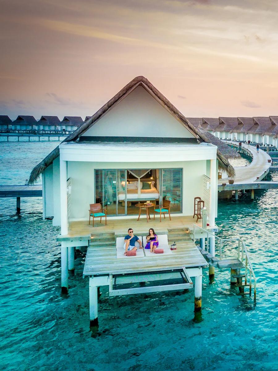 Overwater villas curving out from the island at dusk, a couple relaxing on a private deck in the foreground