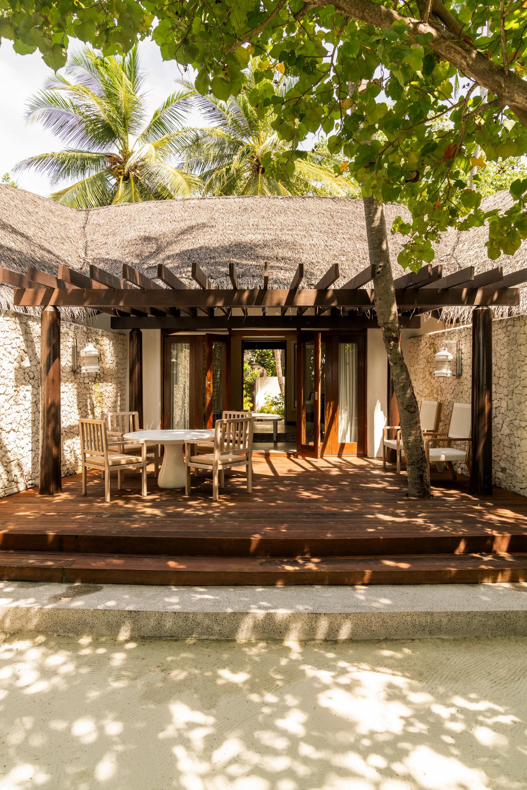 A Beach Villa 2 Bedrooms outdoor terrace with a round dining table and chairs, shaded by a wooden pergola and thatched roof, stone walls framing the space