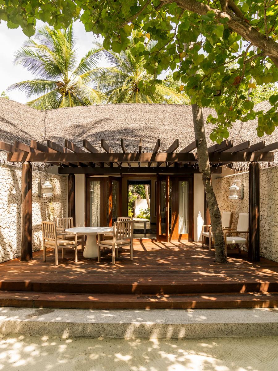 A Beach Villa 2 Bedrooms outdoor terrace with a round dining table and chairs, shaded by a wooden pergola and thatched roof, stone walls framing the space