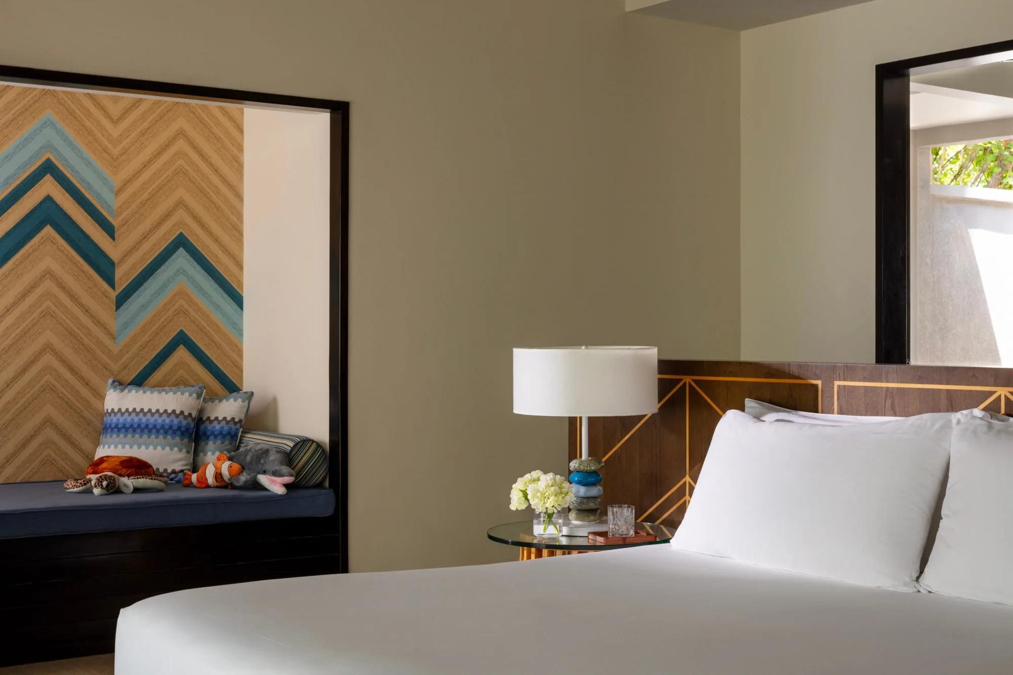 A Beach Villa with Private Pool's window seat scattered with plush turtle, clownfish and dolphin toys, a patterned chevron wall panel behind and a bedside lamp of stacked stone discs
