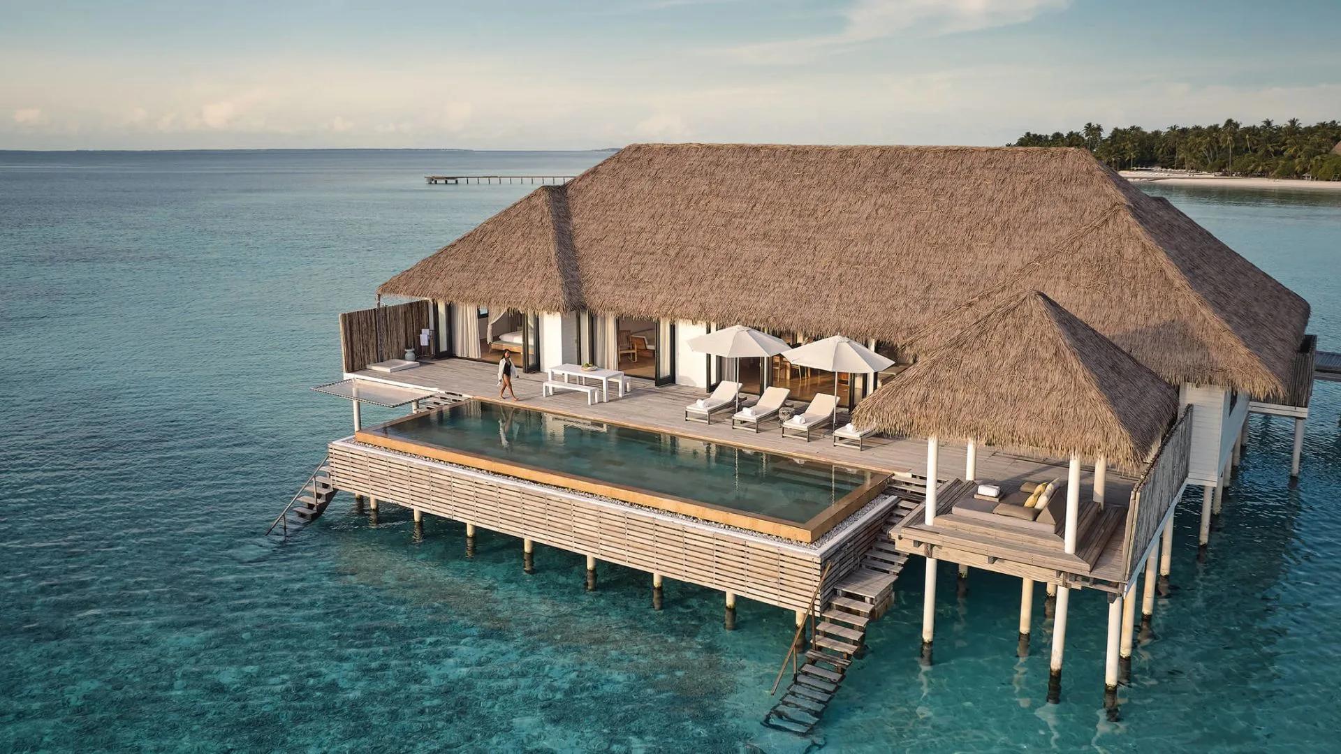 Elevated view of the Maalifushi Water Villa's thatched roof and long pool over the lagoon, with a guest walking the deck and the island visible beyond